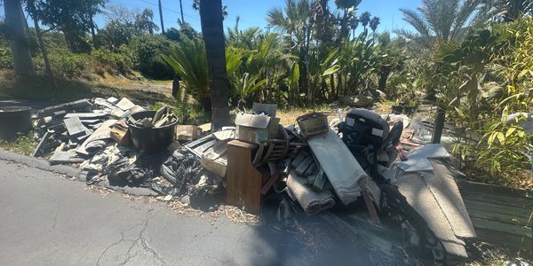 Pile of discarded household items and debris on the roadside under clear blue sky.