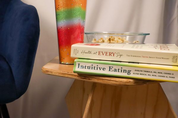 two books, a rainbow water cup and bowl of popcorn on a side table. Intuitive Eating and Health at E