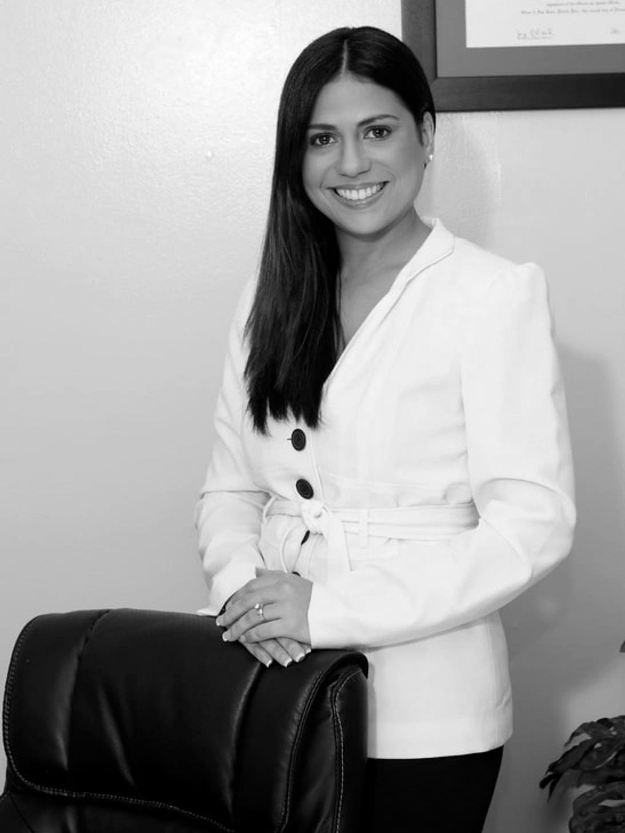 Professional woman in white blazer smiling, standing behind a leather chair.