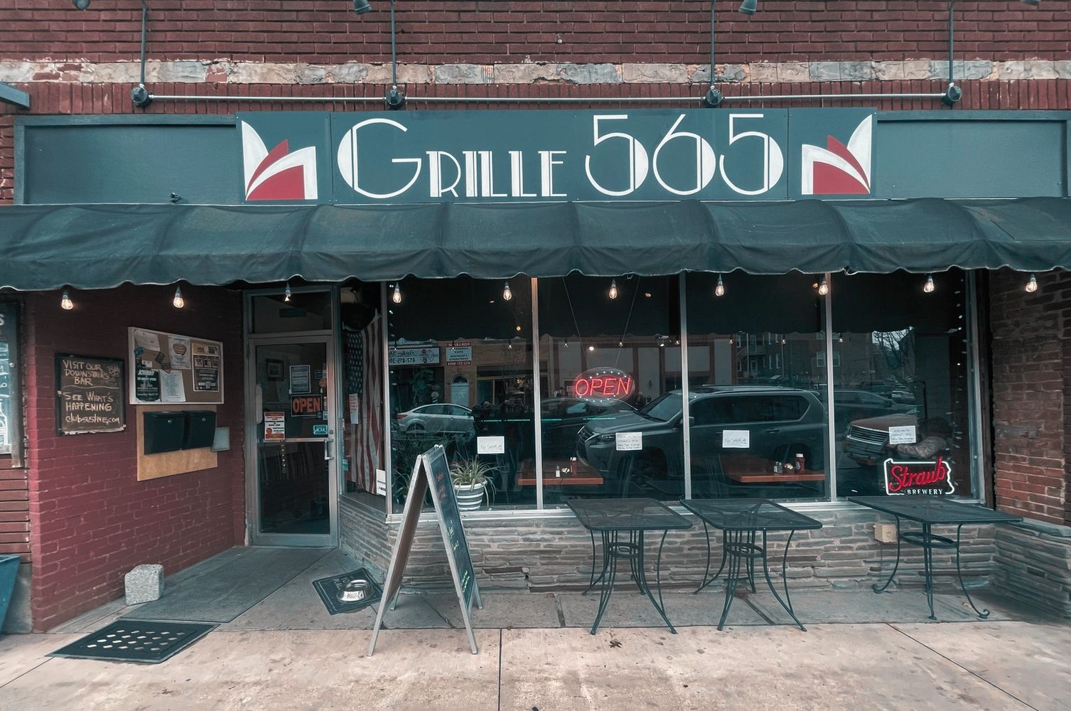 Storefront image, red brick building with a black awning. Sidewalk with outdoor dining tables.