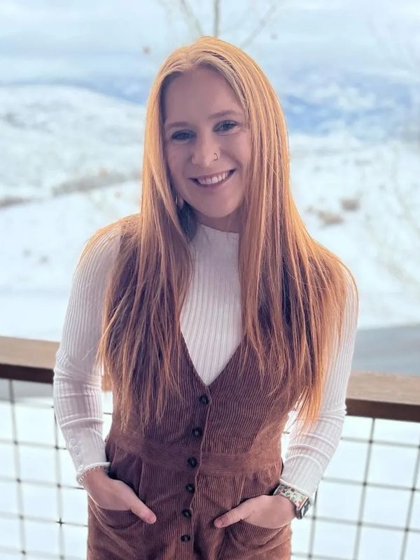 Smiling woman with long red hair in layered winter outfit stands outdoors.