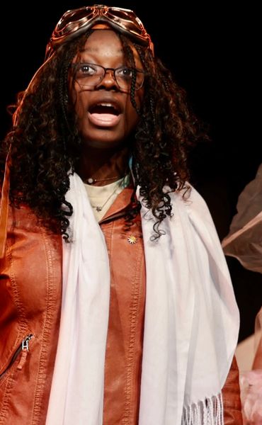 Woman in leather jacket and goggles speaking or performing on stage.
