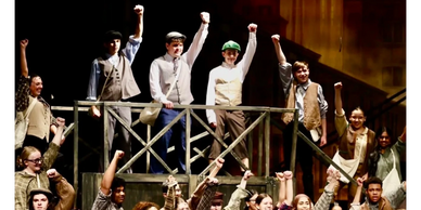 Cast of young actors performing energetically on stage with raised fists.