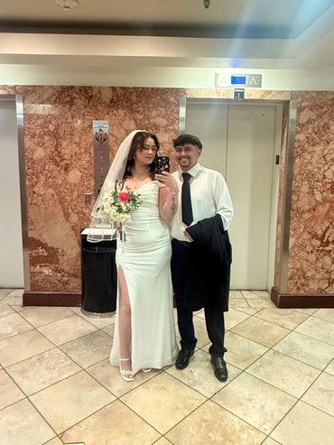 Couple dressed for a wedding, posing in front of an elevator mirror.