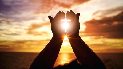 Hands holding a glowing cross against a sunset over water.