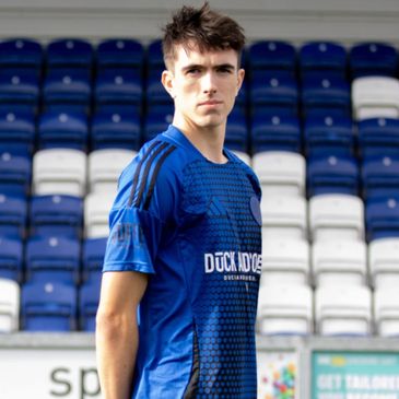Young male soccer player in blue jersey standing in stadium seating area.