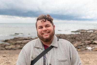 A man in a gray jacket smiles o the beach.