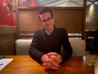 Man sitting at a wooden table in a cozy restaurant with folded hands.