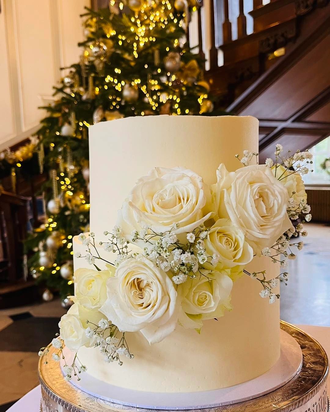 Elegant white cake with roses and Christmas tree in the background.