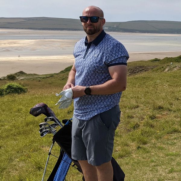 Man in sunglasses holding golf glove by a golf bag on a grassy course near a sandy beach.
