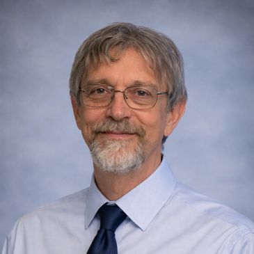 Smiling man with glasses and a beard wearing a tie and light blue shirt.