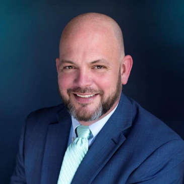 Smiling bald man in a blue suit and green tie against a blue background.