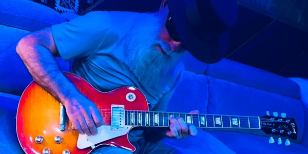 Bearded man in a hat playing an electric guitar on a blue-lit couch.