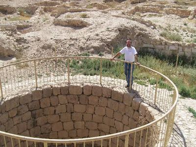 Man standing by a circular stone well with a metal railing in a rocky outdoor area.
