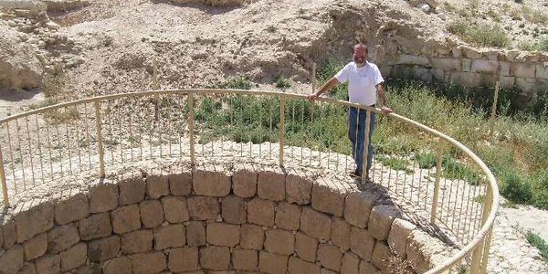 Man standing by a circular stone well with a metal railing in a dry, rocky area.