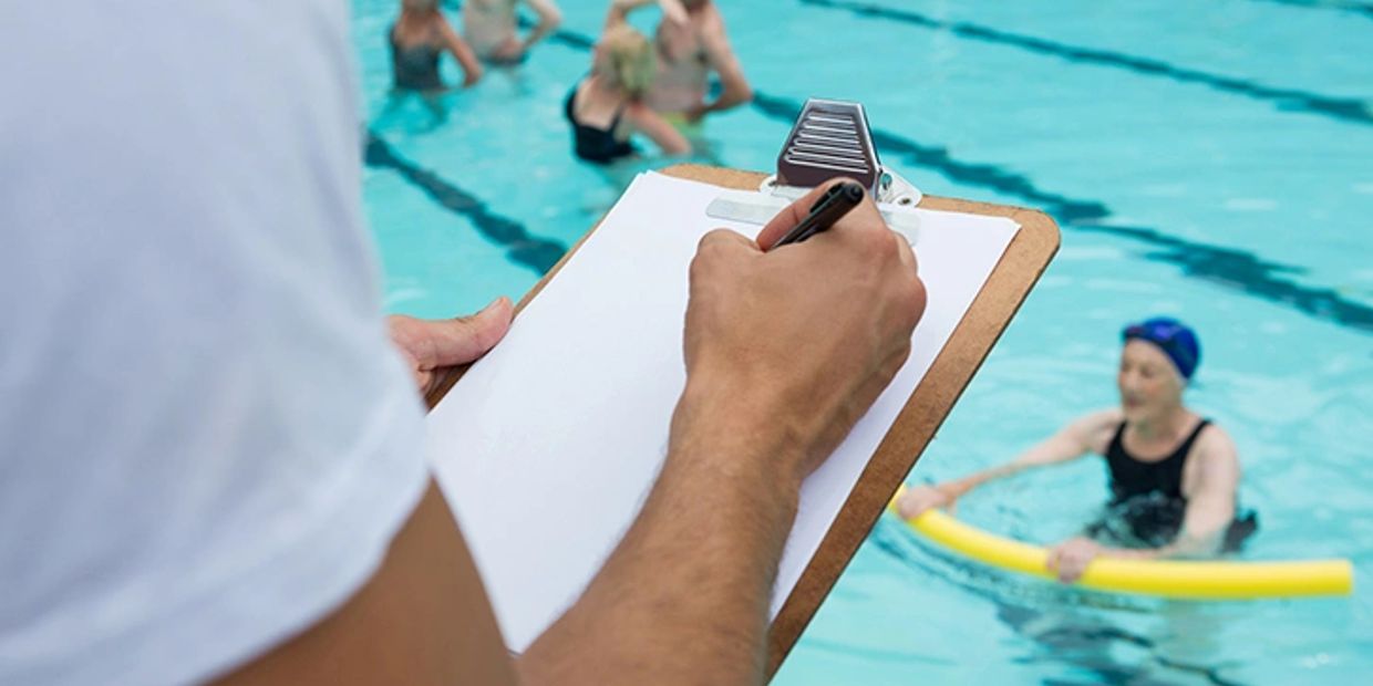 Person with clipboard beside a swimming pool with swimmers.