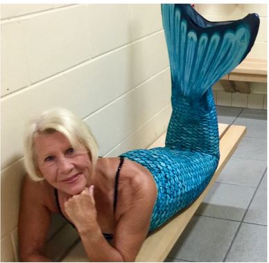 A woman wearing a blue mermaid tail costume, lying on a bench and smiling.