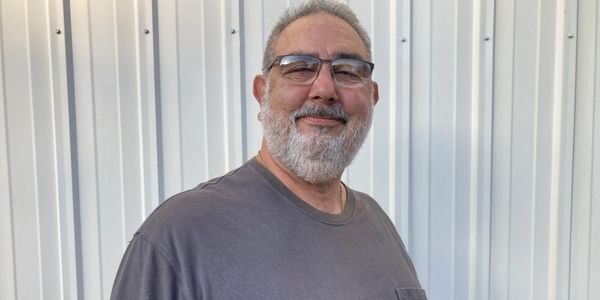 A smiling man with glasses and a beard stands against a corrugated metal wall.