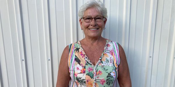 Smiling elderly woman in a colorful floral dress standing against a white wall.