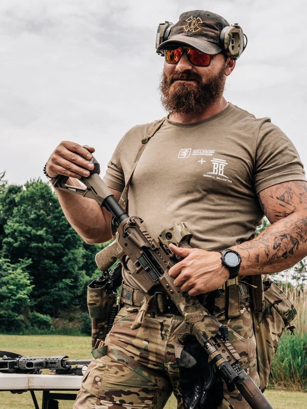 Bearded man in tactical gear holding a rifle outdoors.