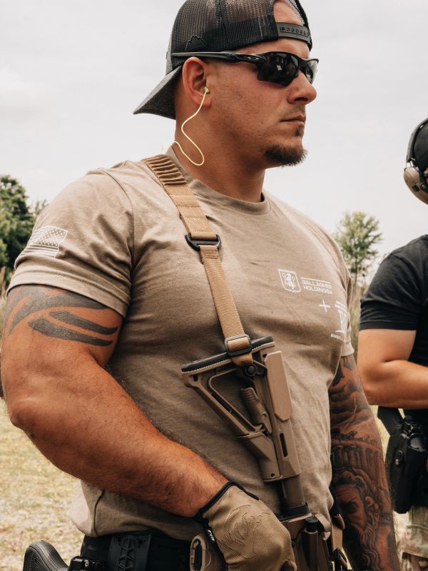 Man in tactical gear holding a rifle, wearing sunglasses and gloves.