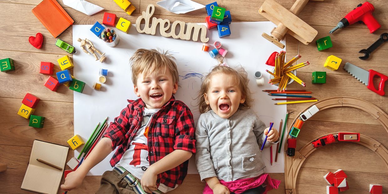 Two happy children lying on the floor surrounded by colorful toys and art supplies.