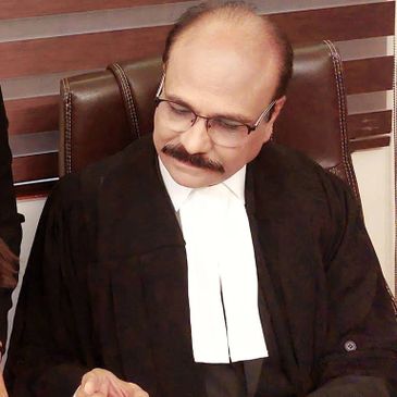 Man in judicial robe and white neckband sits thoughtfully in chair.