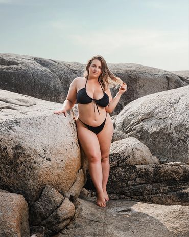 lifestyle photography of a curvy plus size woman wearing swimsuit on rocky beach 