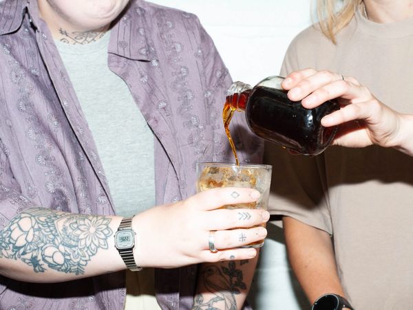 A young woman pours a cold brew into a glass held by another, heavily tattooed, individual.