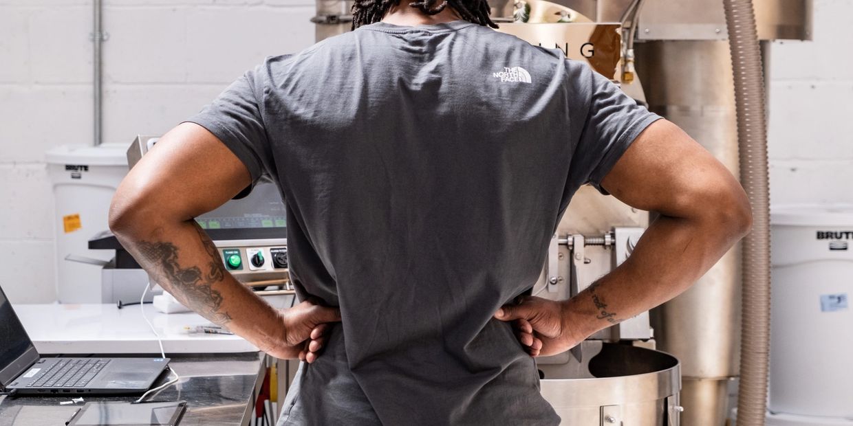 A muscular ban with his back turned and hands on hips looking at a large coffee roaster.