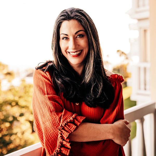 Smiling woman in a red blouse standing on a balcony with arms crossed.