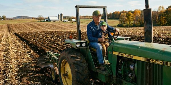 An older man and a child on a John Deere tractor in a farm field.
