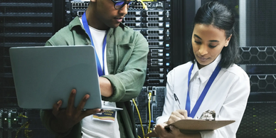 Two IT professionals working in a server room with laptop and clipboard.