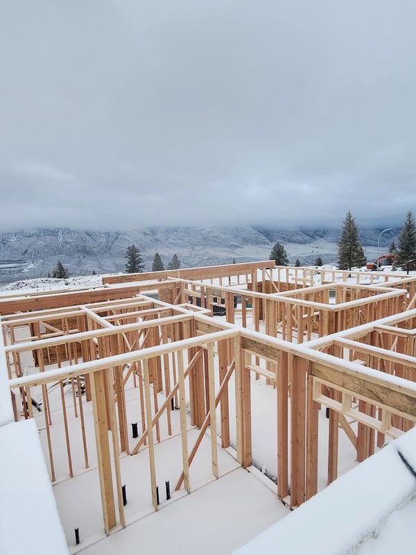 Snow covers a wooden house frame under a cloudy sky in a mountainous area.