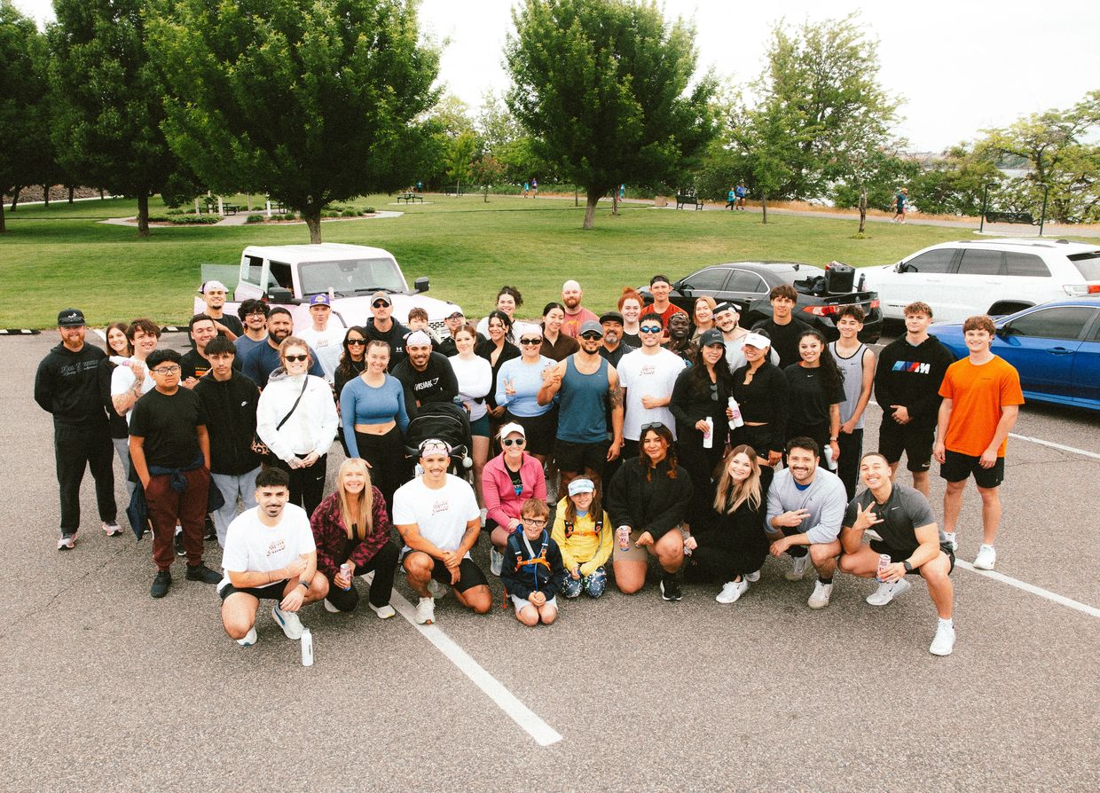 A large group of people posing together outdoors in a parking lot with trees and cars in the background.