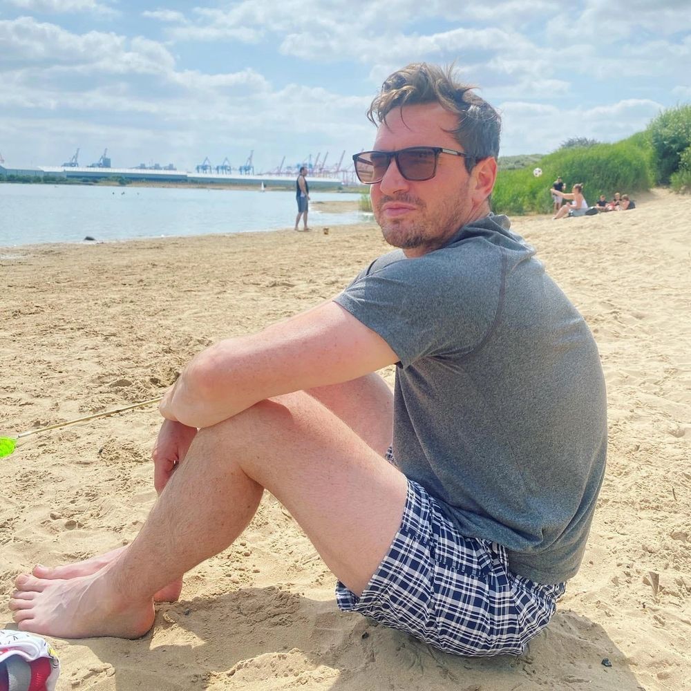 A photo of Sam. He is sat on the beach and is smiling at the camera.