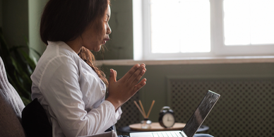 Woman praying while sitting with a laptop in front of a window.