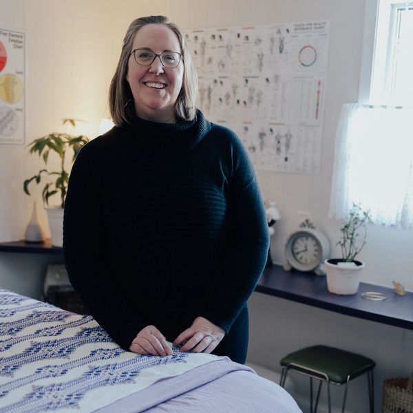 Ruth stands in her office: clean, comfortable, homey. Touch for Health charts visible on the wall. M