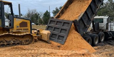 Dump truck unloading sand near a bulldozer at a construction site.