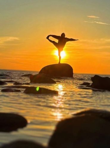Silhouette of person doing yoga on rock at sunset.