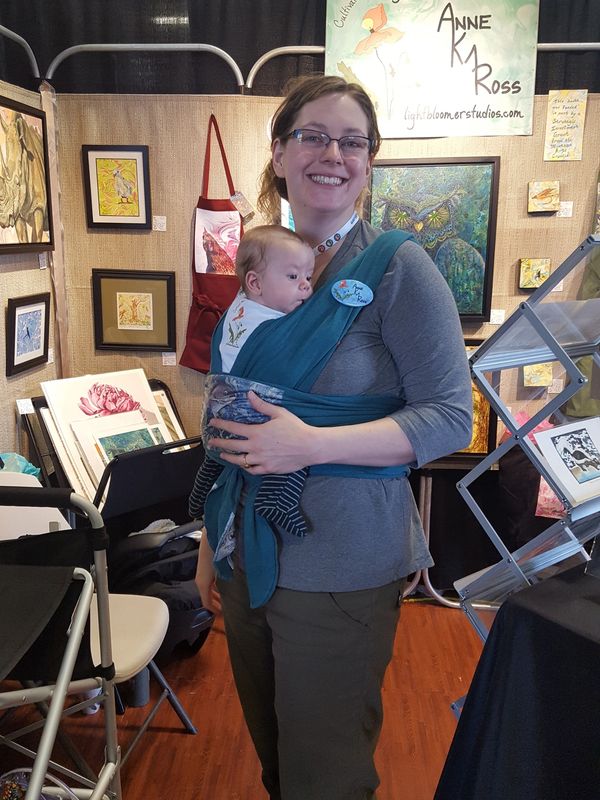 Anne standing in her art show booth with her infant in a wrap carrier on her chest.