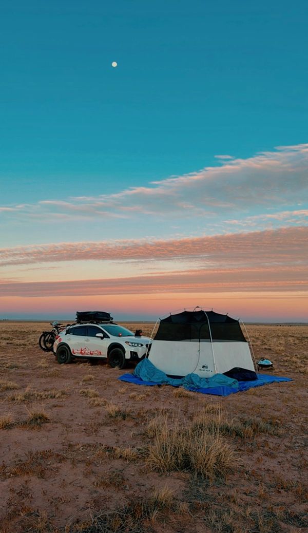 A car and tent set up in a dry field under a moonlit sky at sunset.