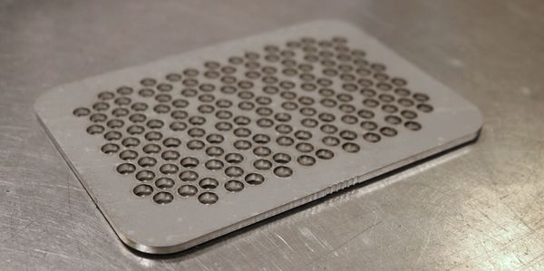 A rectangular, metal bulk head with small holes cut in it. The part is sitting on a metal table. 