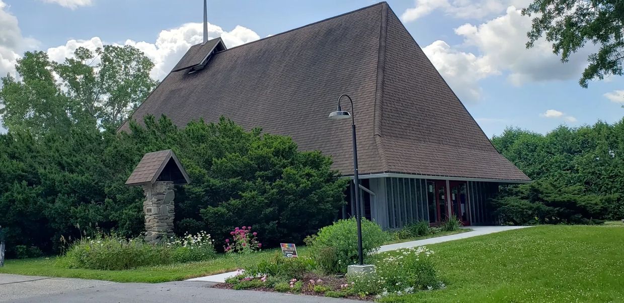 A church with a steep roof and surrounding greenery on a sunny day.