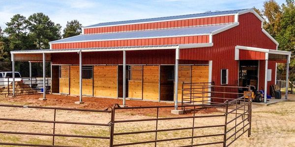 Red barn with wooden stalls and fenced area under clear sky.