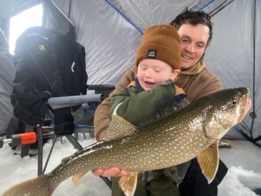 A father and child proudly hold a large fish inside an ice fishing tent.