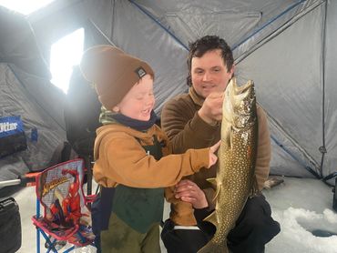 A man and child joyfully display a large fish inside an ice fishing tent.