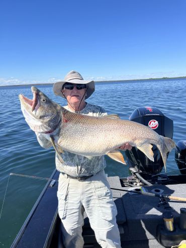 Fisherman holding a large fish on a boat under clear blue sky.