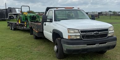 White Chevrolet truck towing a trailer with a green tractor under cloudy skies.