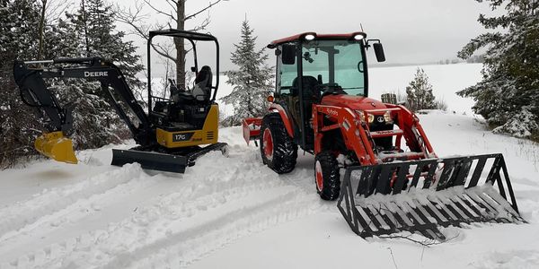 Two tractors parked on snow-covered ground surrounded by trees.